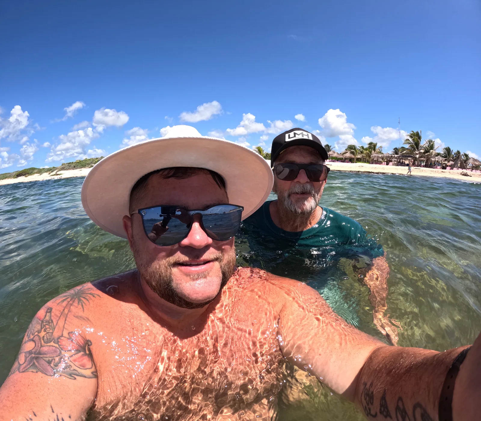Chris and Randy, the son and father who make Salt of the Page, taking a selfie waist-deep in clear turquoise water, with a white beach, palm trees and a blue sky behind them.